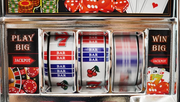 A close-up view of a slot machine with vibrant colors and various gambling symbols such as dice, coins, and playing cards. The reels display a combination of symbols, including bars, cherries, and the number seven. The chrome exterior and colorful details convey the theme of casino and gambling.