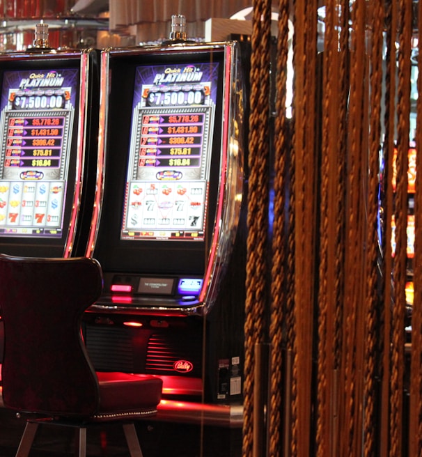 A pair of casino slot machines, each with detailed screens displaying various game outcomes and jackpot amounts. The chairs in front of the slot machines are dark and cushioned for comfort. The background features a hanging curtain made of golden ropes and other casino decorations are visible.
