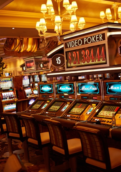 A casino floor featuring rows of brightly lit video poker machines. The area is illuminated by elegant chandeliers, with a display board showing a large sum of money, suggesting a possible jackpot. The rich decor includes luxurious carpets and comfortable seating, adding to the opulent atmosphere.