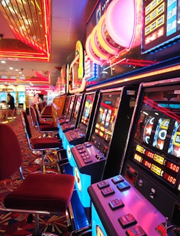 A brightly lit casino interior featuring a row of colorful slot machines. The neon lights create a vibrant atmosphere, with patterned carpeting adding to the decor. Stools are aligned neatly in front of each machine.