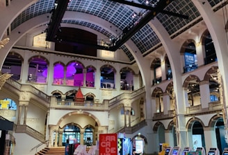 The interior of a large, ornately designed building with arched ceilings and intricate architectural details. The space features gaming machines lined up along the floor, under a high ceiling with a grid pattern. Lighting in purple and white tones enhances the ambiance.