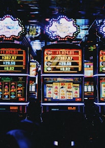 A row of colorful slot machines lit with bright neon lights, showcasing various graphical elements on the screens, indicating different games and jackpots. The atmosphere suggests a lively casino setting, bustling with excitement.