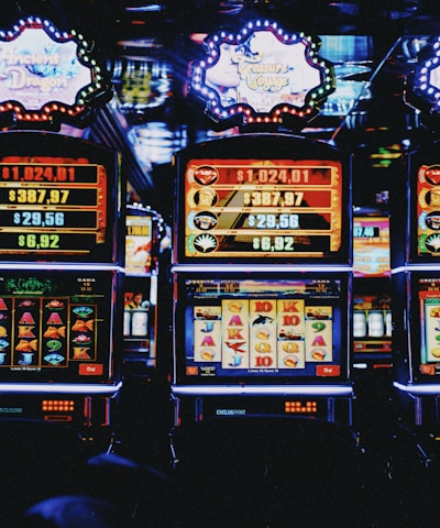A row of colorful slot machines lit with bright neon lights, showcasing various graphical elements on the screens, indicating different games and jackpots. The atmosphere suggests a lively casino setting, bustling with excitement.