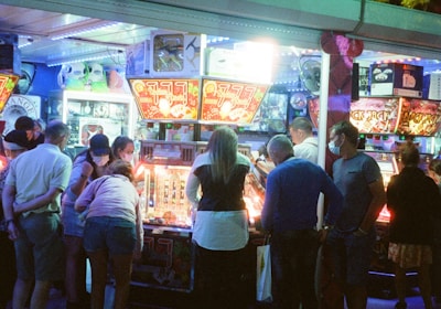 A group of people standing in front of an illuminated amusement arcade, engaging with various gaming machines. The environment is bustling with activity, and colorful lights create a vibrant atmosphere. The ceiling is decorated with large, playful images of dice and other game-related graphics, enhancing the lively mood.
