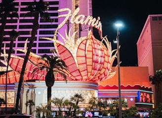 A brightly lit casino with neon lights in vibrant red, pink, and blue, featuring a large flamingo signage. Palm trees are visible in the foreground with vehicles passing by on the street at night.