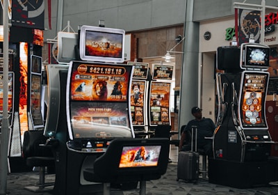 A collection of slot machines is situated inside a building with bright, colorful screens displaying various games and gaming themes. A person is seated nearby, seemingly relaxed with luggage beside them, and the atmosphere is lively with electronic displays.
